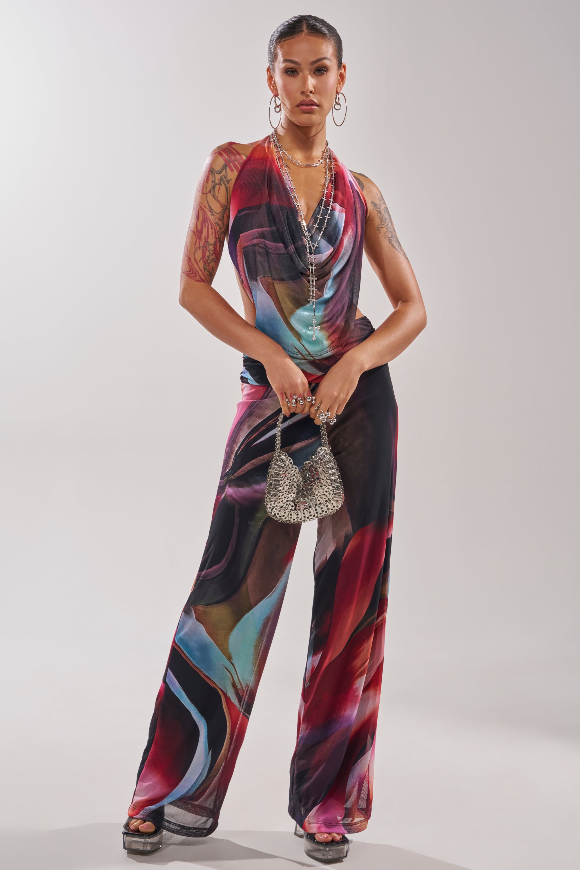 A woman stands confidently in the MAIN CHARACTER ENERGY COWL NECK JUMPSUIT, paired with platform sandals, layered necklaces, large hoop earrings, and a silver beaded handbag against a plain light background. 2