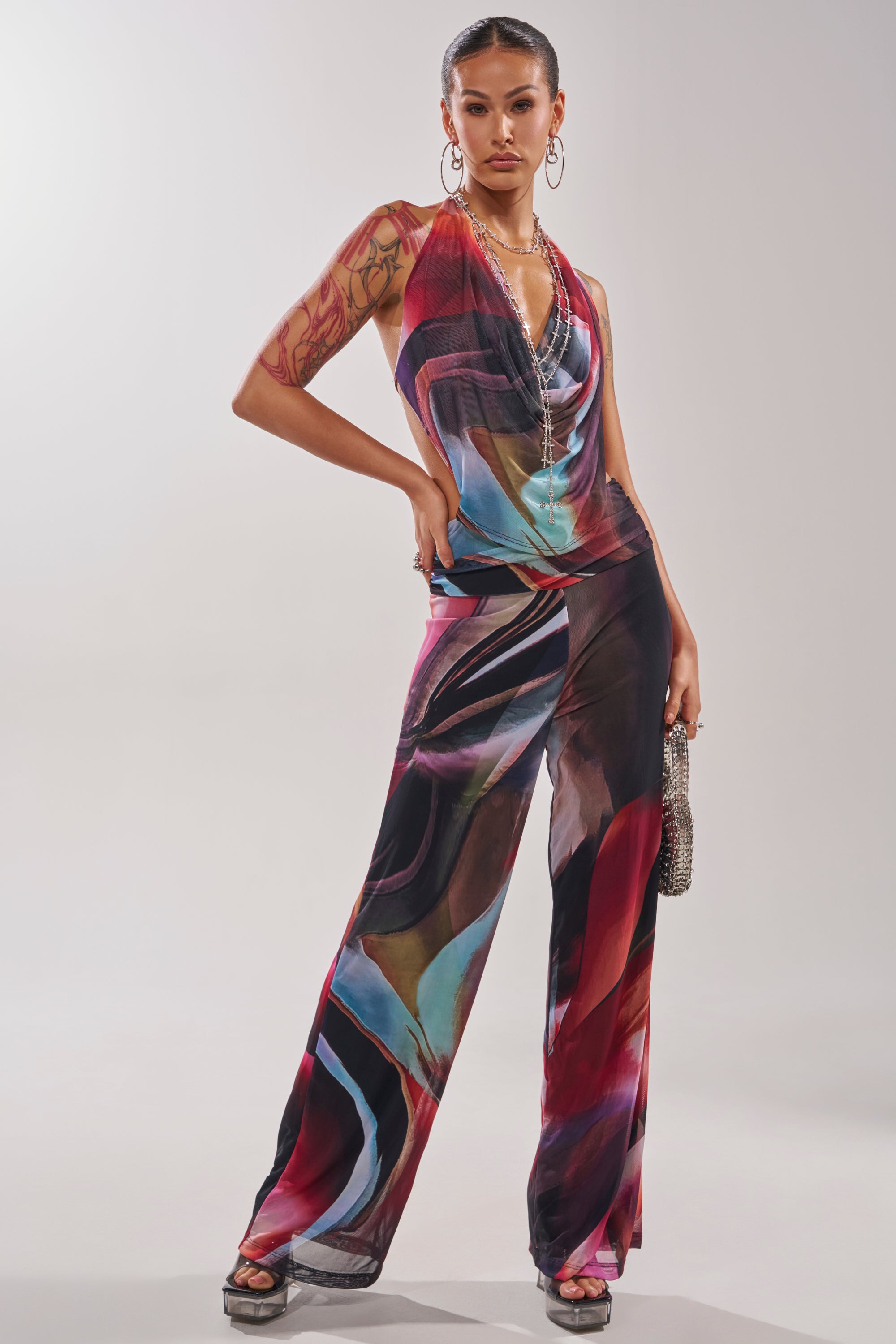 A woman poses confidently in the MAIN CHARACTER ENERGY COWL NECK JUMPSUIT, featuring a multicolor abstract swirl print. She pairs it with hoop earrings, layered necklaces, and a textured handbag for a chic statement look. 3