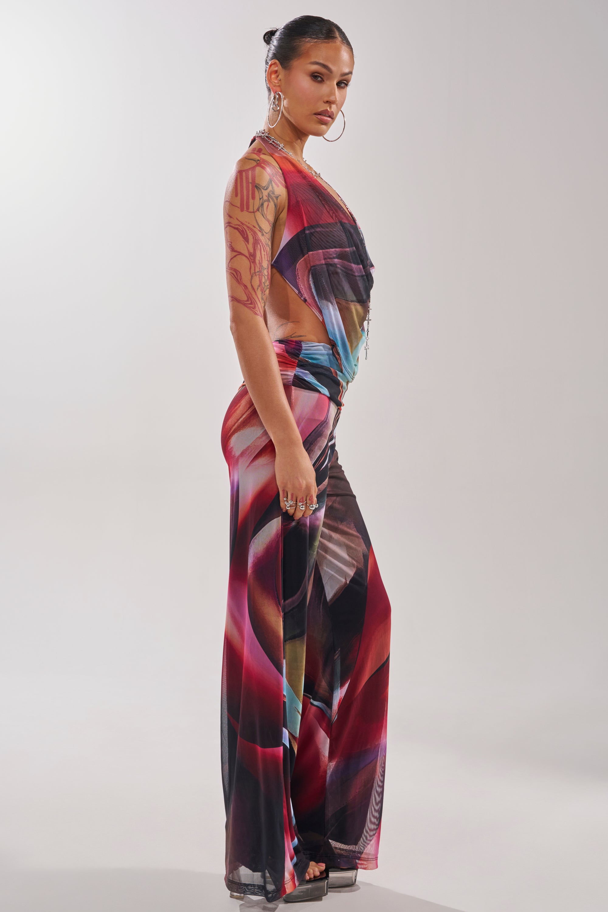 Woman with dark hair in a bun, large hoop earrings, and tattoos on her upper arm models the MAIN CHARACTER ENERGY COWL NECK JUMPSUIT featuring a multicolor swirl print. She stands sideways against a plain background. 4