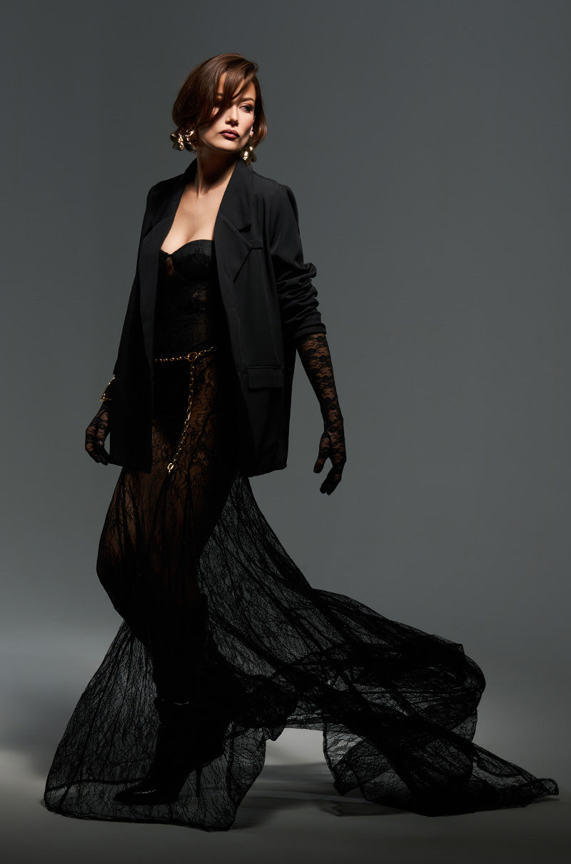 Wearing the TROUBLES HERE LACE MAXI DRESS, someone poses elegantly in a flowing black lace maxi dress with sheer, stretchy details, paired with a dark jacket and gloves. The curve-skimming silhouette is beautifully enhanced by gold earrings and a waist chain, set against a gray background.