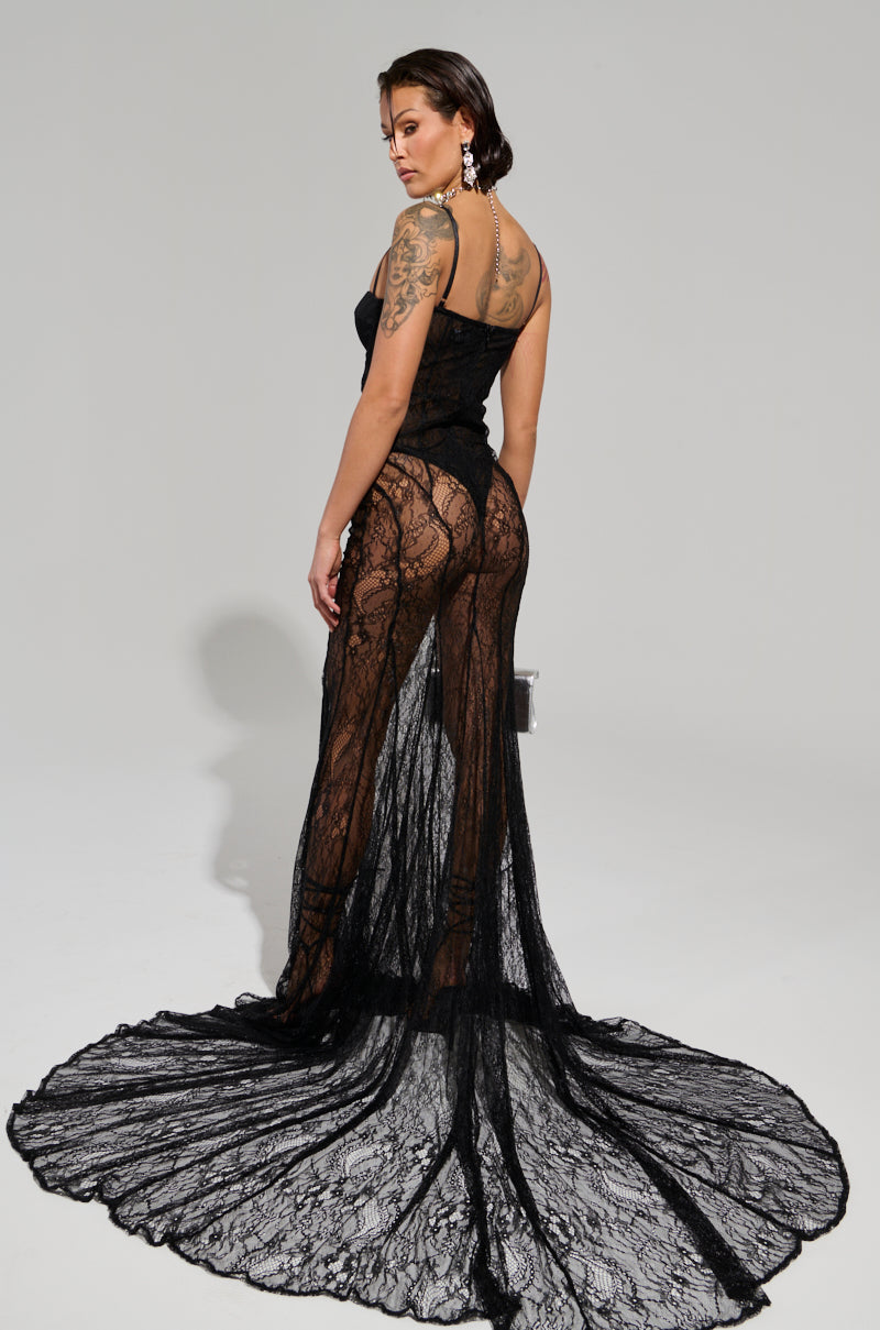 A model poses in the TROUBLES HERE LACE MAXI DRESS, a sheer and stretchy lace maxi dress featuring a long, flowing train. Her back showcases tattoos, and she accessorizes with large, ornate earrings that enhance the curve-skimming silhouette. The background is a simple light gray.