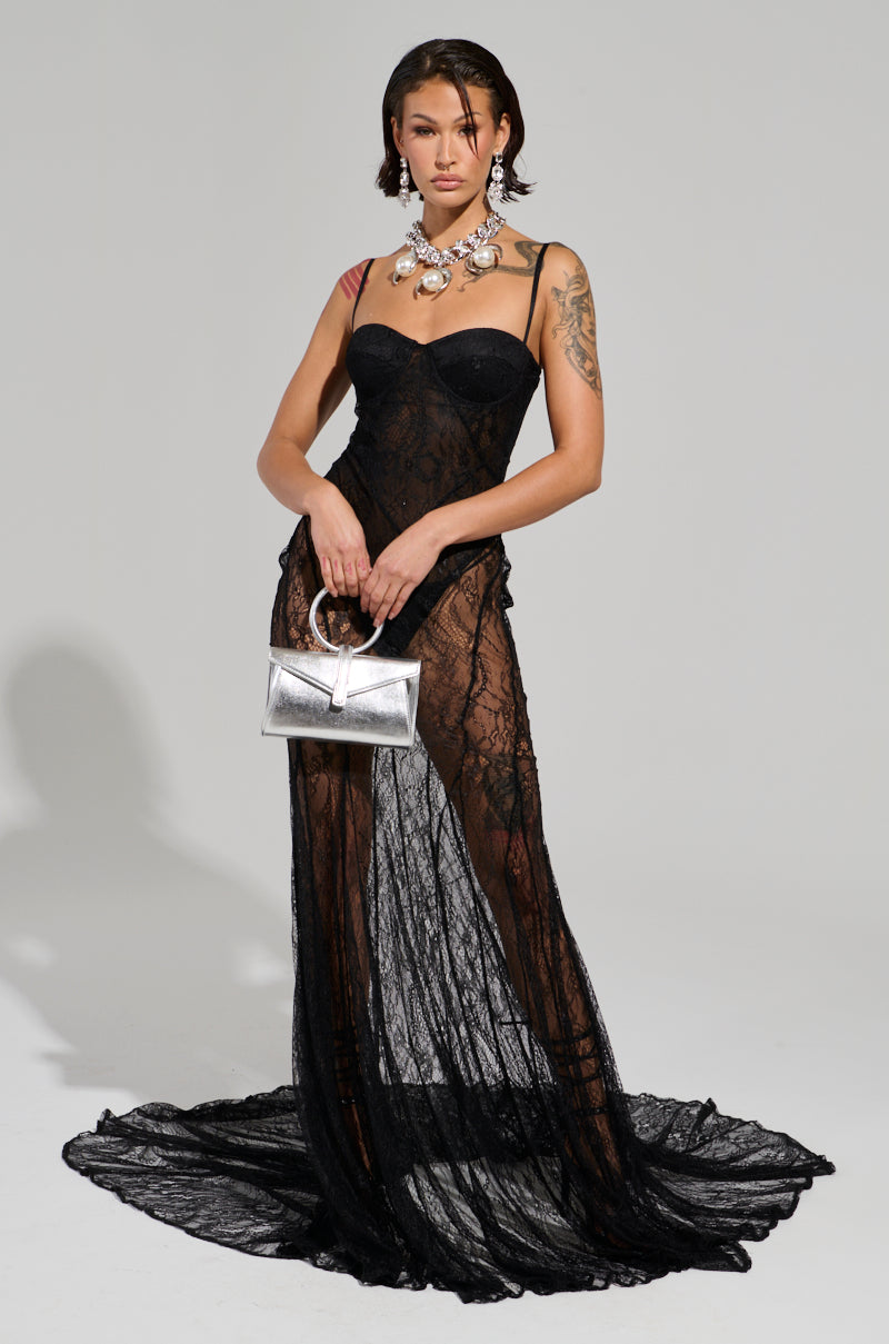 A person showcases a sleek silhouette in the TROUBLES HERE LACE MAXI DRESS, made from sheer stretchy lace, against a plain background. They carry a metallic silver handbag and accessorize with statement jewelry, including a chunky necklace. A tattoo is visible on their upper arm, contributing an edgy touch to the elegant outfit.