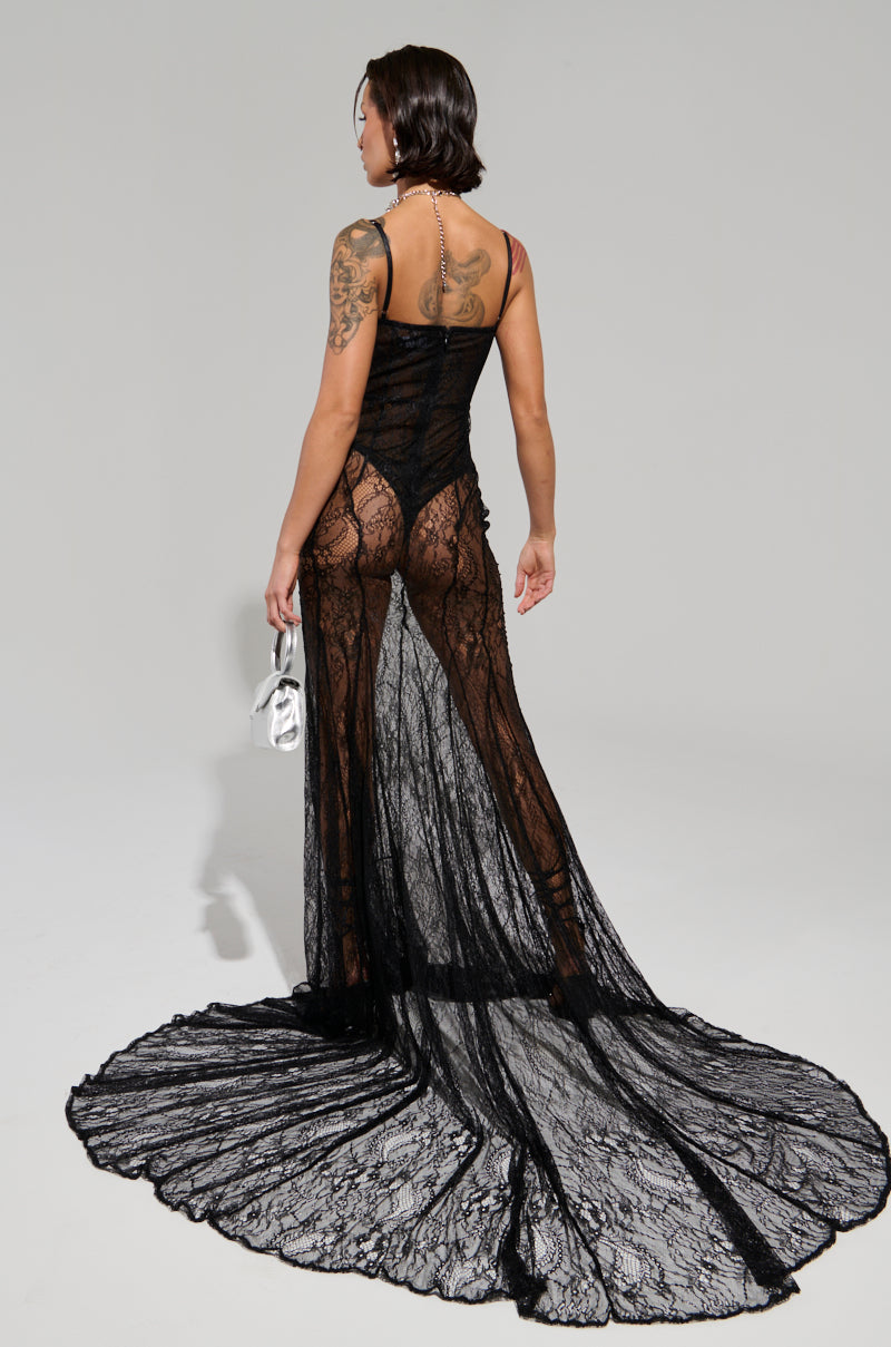 A person with tattoos models the TROUBLES HERE LACE MAXI DRESS, which features a curve-skimming silhouette of sheer stretchy lace that flows into a train. They hold white strappy high-heeled shoes while facing away from the camera against a plain, light-colored backdrop.