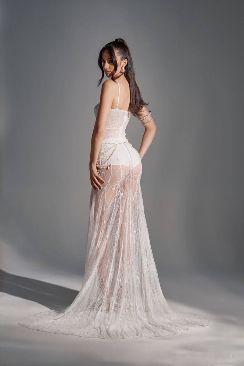 A woman stands on a gray background in the TROUBLES HERE LACE MAXI DRESS IN WHITE, featuring sheer stretchy lace, a fitted bodice, train, and an elegant sweetheart neckline. She looks back over her shoulder with a half-up hairstyle.