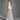 A woman stands on a gray background in the TROUBLES HERE LACE MAXI DRESS IN WHITE, featuring sheer stretchy lace, a fitted bodice, train, and an elegant sweetheart neckline. She looks back over her shoulder with a half-up hairstyle.
