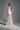 A woman stands on a gray background in the TROUBLES HERE LACE MAXI DRESS IN WHITE, featuring sheer stretchy lace, a fitted bodice, train, and an elegant sweetheart neckline. She looks back over her shoulder with a half-up hairstyle.