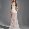 A woman stands on a gray background in the TROUBLES HERE LACE MAXI DRESS IN WHITE, featuring sheer stretchy lace, a fitted bodice, train, and an elegant sweetheart neckline. She looks back over her shoulder with a half-up hairstyle.