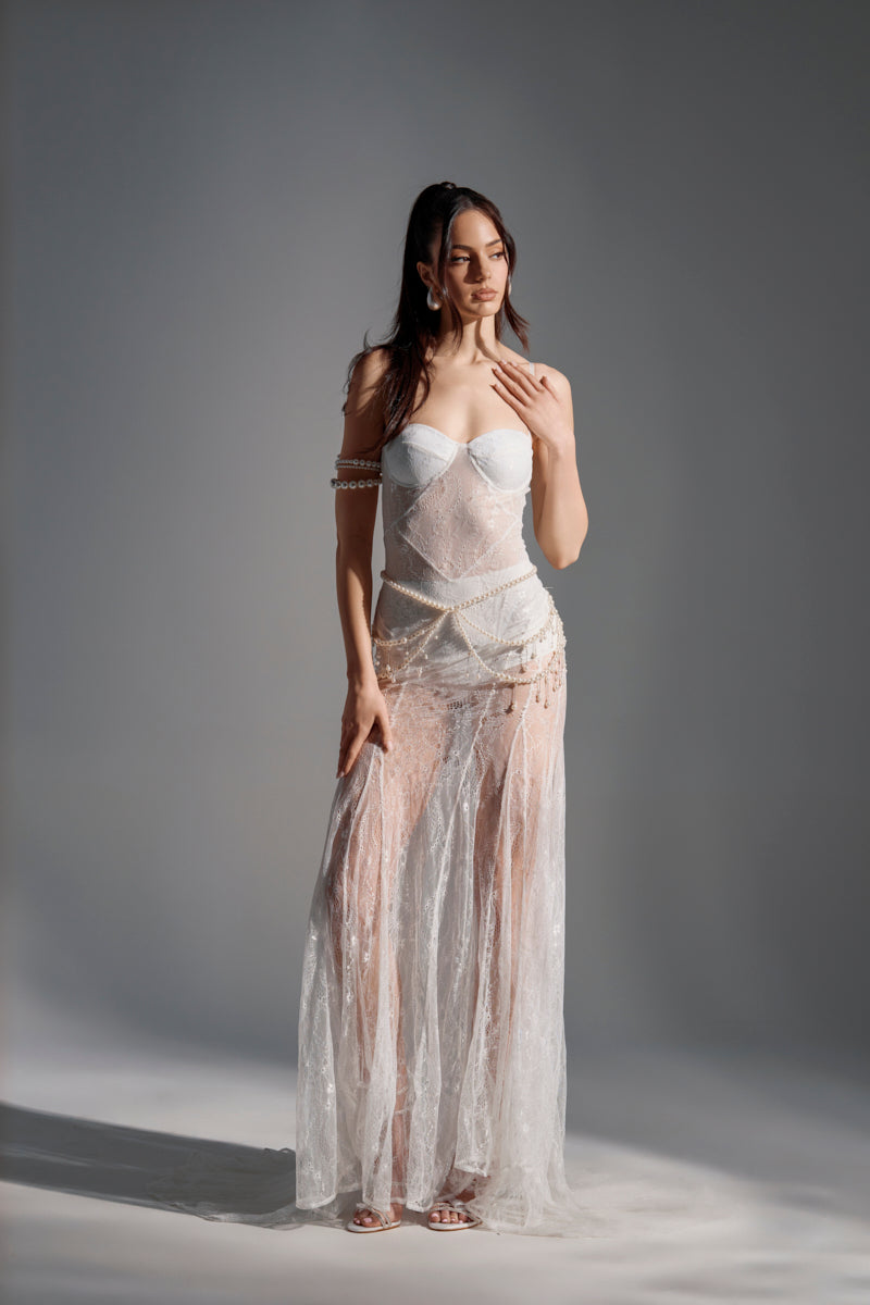 A woman poses in the TROUBLES HERE LACE MAXI DRESS IN WHITE, featuring a sheer lace design, sweetheart neckline, pearl details, and strapless bodice. Her long brown hair flows over her shoulders as she stands against a plain gray background.