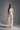 A woman poses in the TROUBLES HERE LACE MAXI DRESS IN WHITE, featuring a sheer lace design, sweetheart neckline, pearl details, and strapless bodice. Her long brown hair flows over her shoulders as she stands against a plain gray background.