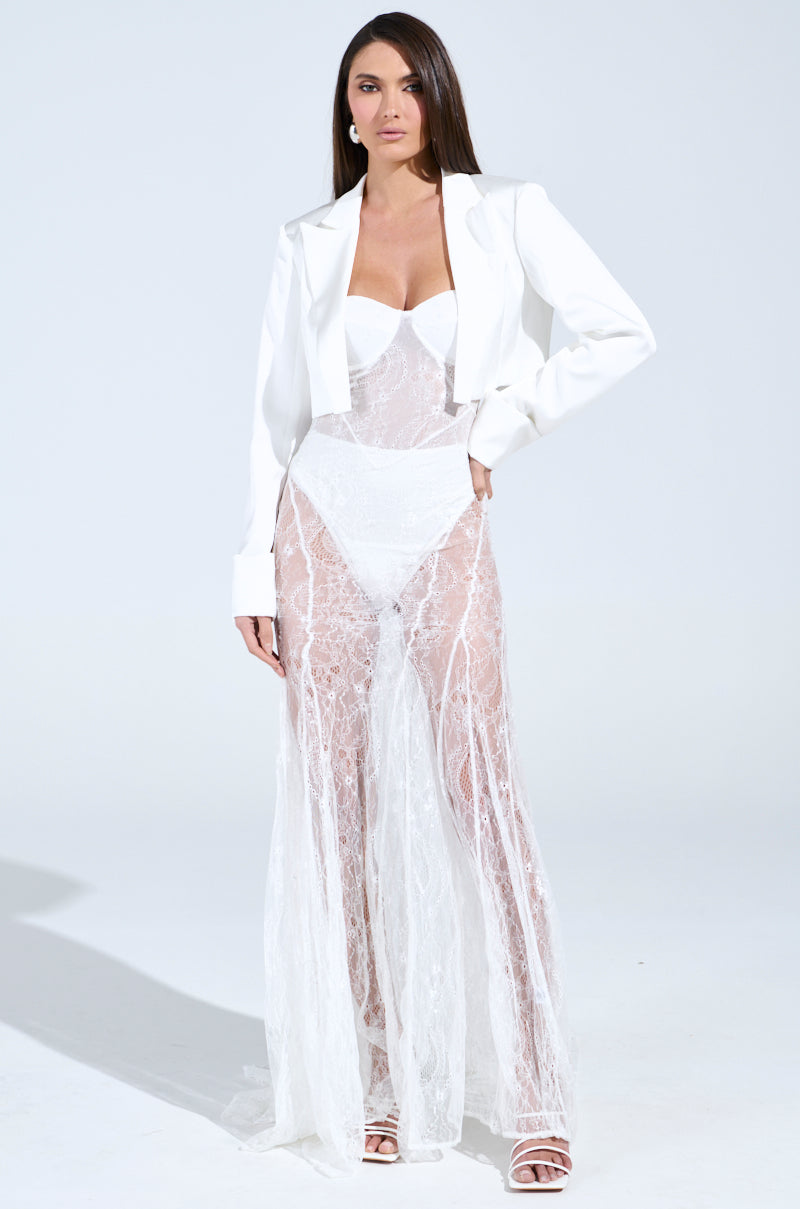 Wearing the TROUBLES HERE LACE MAXI DRESS IN WHITE, a person confidently poses against a plain backdrop. Their long hair cascades over the sheer fabric, enhancing the elegant and poised appearance of the ensemble's sweetheart neckline with high-waisted undergarments and a structured cropped blazer.