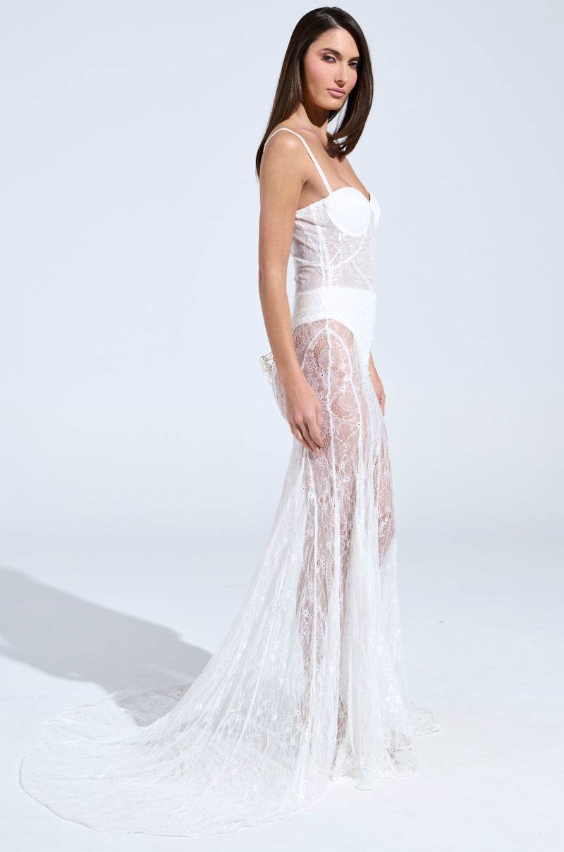 A woman poses in the TROUBLES HERE LACE MAXI DRESS IN WHITE. This sheer spaghetti-strap maxi dress features a sweetheart neckline and semi-transparent design, spotlighting her side profile on a plain white background with arms relaxed by her sides.
