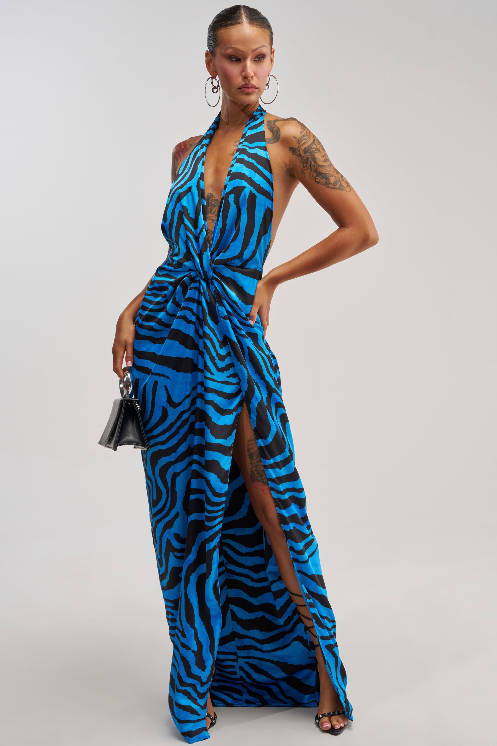 A woman poses in the CRAZY FOR YOU BLUE ZEBRA MULTI MAXI DRESS, a sleeveless satin halter maxi with a high slit, accessorized with a small black handbag, hoop earrings, and black sandals.