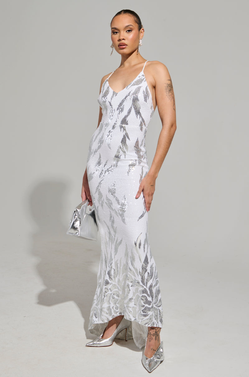 A person poses confidently in the WINTER WHITES SEQUIN MAXI DRESS, showcasing its shimmering silver and white sequins and bodycon silhouette. They hold a matching silver clutch and wear metallic heels, radiating elegance against a simple, neutral background.