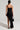 A woman stands with her back to the camera, showcasing the NEW BOO ASYMMETRICAL MAXI DRESS, a long black dress featuring a thigh-high slit. Paired with strappy heels and complemented by a black handbag, her hair is styled in a low ponytail against a simple, light-colored backdrop.