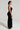 A woman stands in profile facing left, highlighting the sleek design of the NEW BOO ASYMMETRICAL MAXI DRESS, featuring cut-out details. Her hair is styled up and she wears large hoop earrings while holding a small black clutch against a plain, light-colored background.