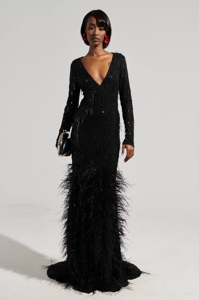 A woman stands against a plain background, wearing the YOURS TRULY Feather Sequin Embellished Maxi Dress. This elegant black dress features a plunging V-neckline and a curve-hugging bodice that flows into feathered details at the bottom. She accessorizes with a black clutch and large red earrings.