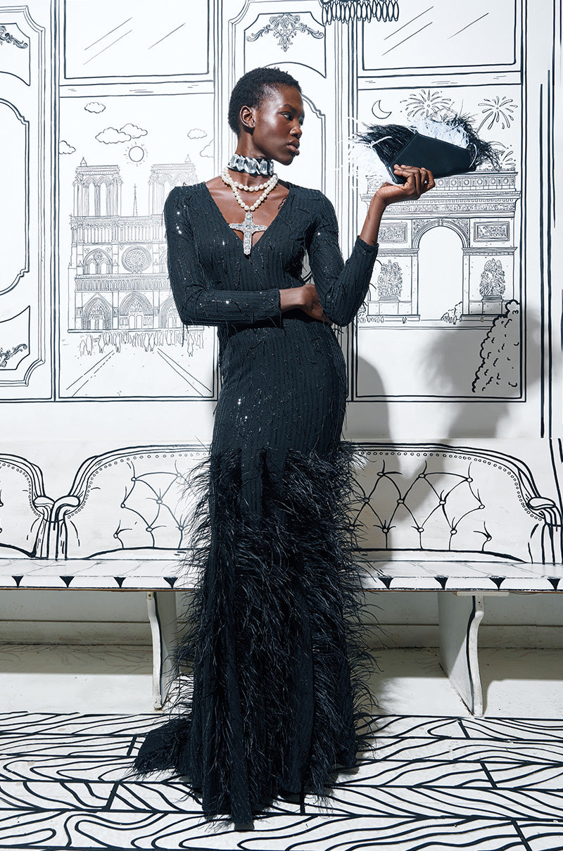 A person wearing the YOURS TRULY FEATHER SEQUIN EMBELLISHED MAXI DRESS, which showcases a plunging V-neckline and curve-hugging bodice, poses indoors. Their hand is adorned with a feathered accessory, set against a backdrop featuring a sketch-like wall design with architectural elements.