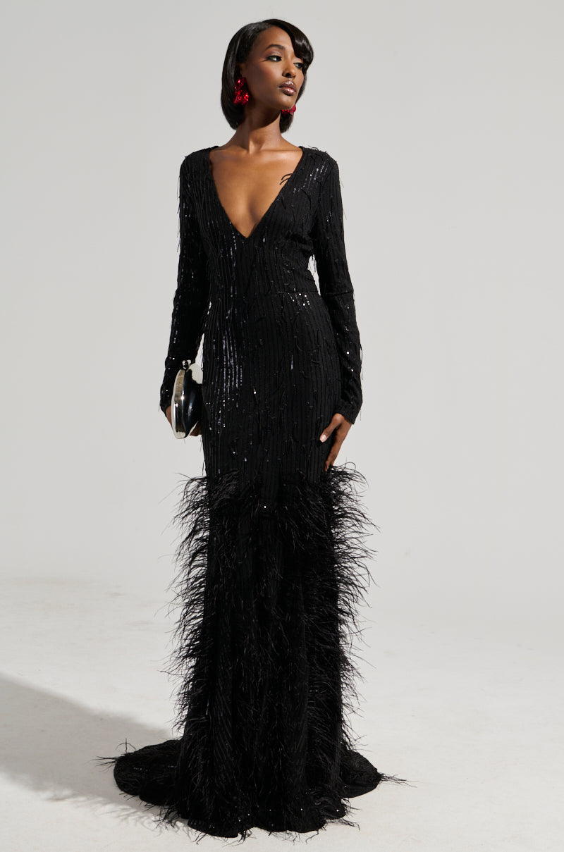 A woman stands confidently in the YOURS TRULY Feather Sequin Embellished Maxi Dress, its curve-hugging bodice accentuating her silhouette. The plunging V-neckline adds an elegant touch. She holds a black clutch and wears large red earrings against a simple, neutral background.
