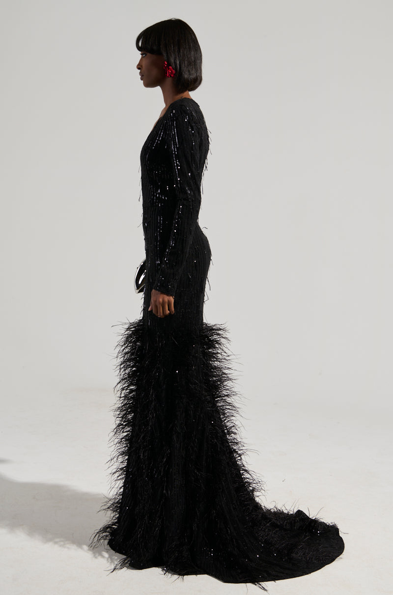 A woman stands in profile, showcasing the YOURS TRULY Feather Sequin Embellished Maxi Dress with a plunging V-neckline. Her floor-length black gown features a curve-hugging bodice adorned with sequins and feathers at the bottom. She has short hair and wears red earrings against a minimal light gray background.