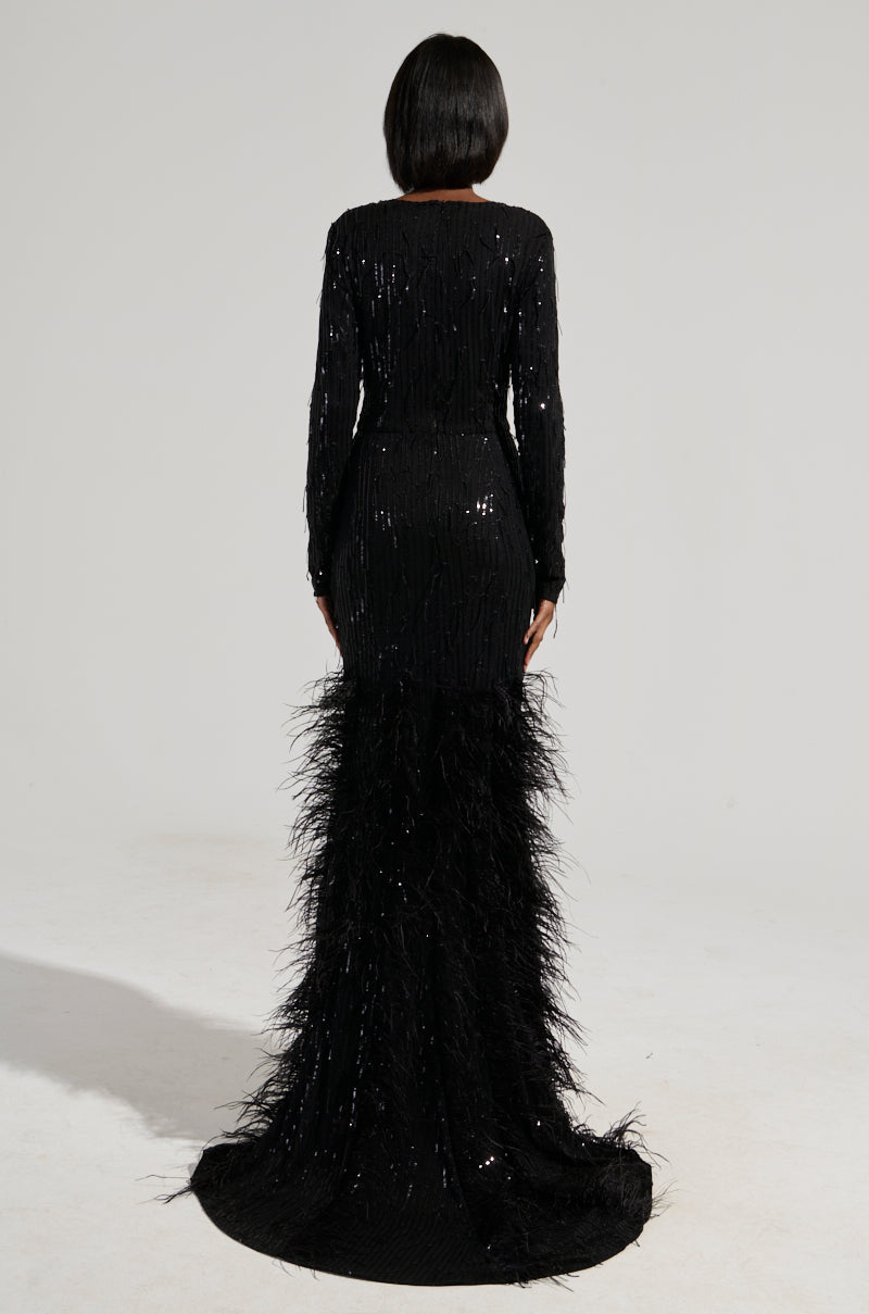 A person with short black hair stands facing away, wearing the YOURS TRULY Feather Sequin Embellished Maxi Dress, featuring feathers that flow to the floor. The curve-hugging bodice and long sleeves create an elegant silhouette against a plain background.