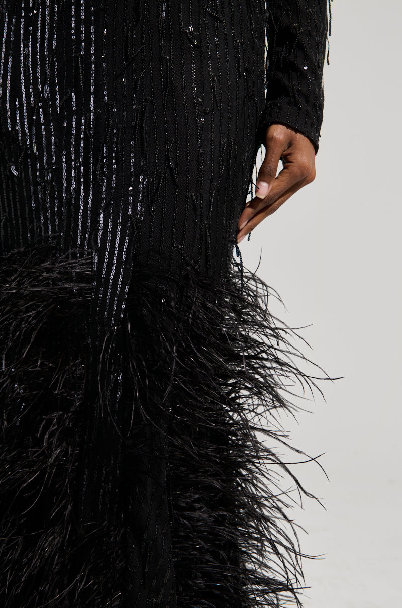A person is adorned in the YOURS TRULY FEATHER SEQUIN EMBELLISHED MAXI DRESS, which boasts a curve-hugging bodice and an elegant, shimmering texture. This long black dress showcases a feathered hem that catches the light beautifully. Their hand lightly touches the side of the plunging V-neckline, enhancing its graceful allure.