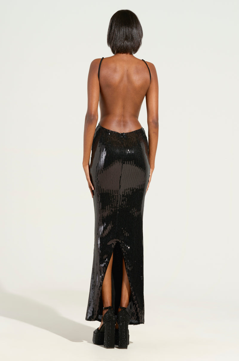 A person with short hair is wearing the ALL NIGHT LONG SEQUIN MAXI DRESS, showcasing a striking open back and delicate thin straps. This floor-length, black dress, featuring a slit at the back, beautifully stands out against the plain light background.