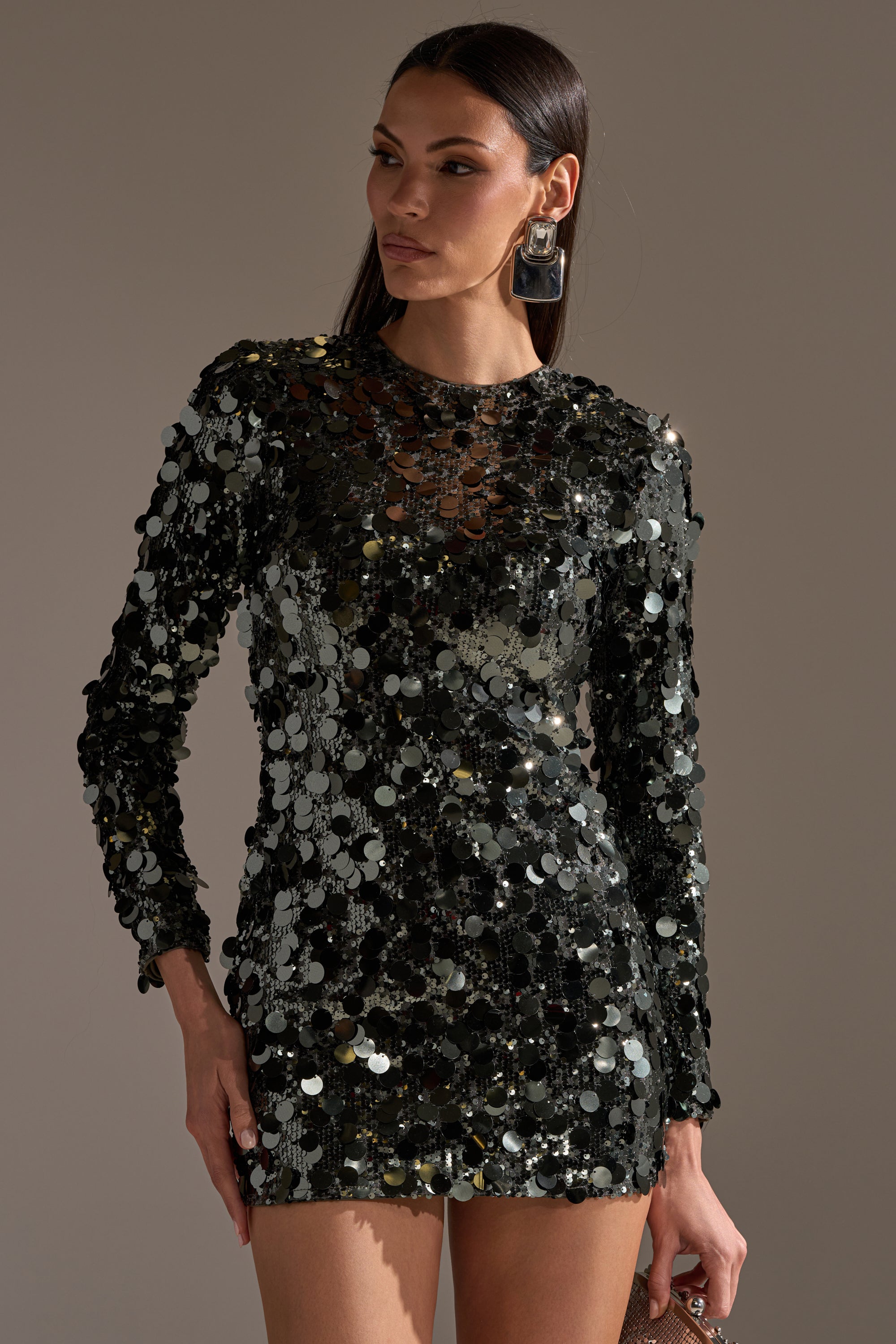 A woman stands against a plain background in the SHE'S ALL THAT SEQUIN MINI DRESS IN OLIVE, featuring long sleeves and shimmering sequins. Her straight hair, statement earrings, and confident expression create the perfect party-ready look.