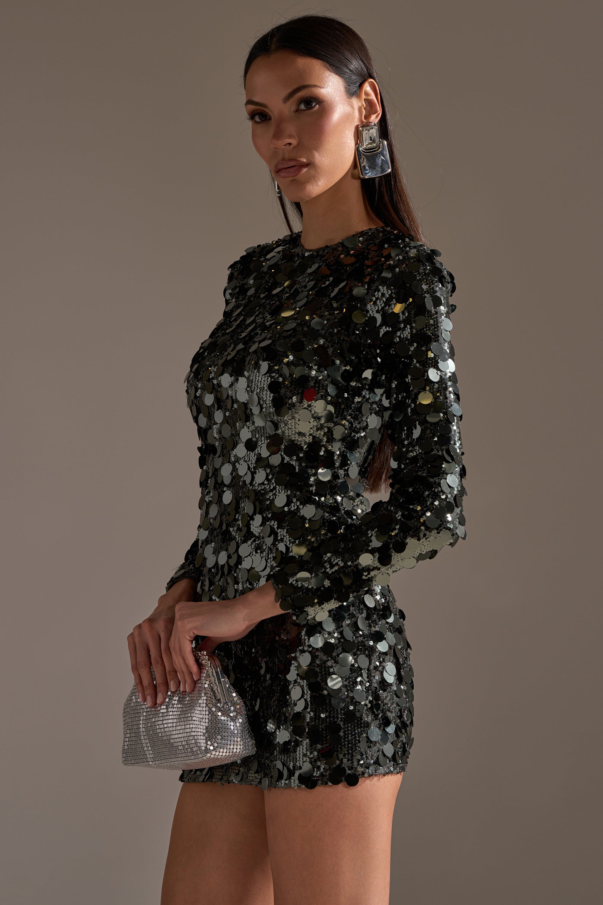 A woman in the SHE'S ALL THAT SEQUIN MINI DRESS IN OLIVE holds a sparkly silver clutch and wears large rectangular earrings, striking a pose against a plain background—the ultimate party dress look.