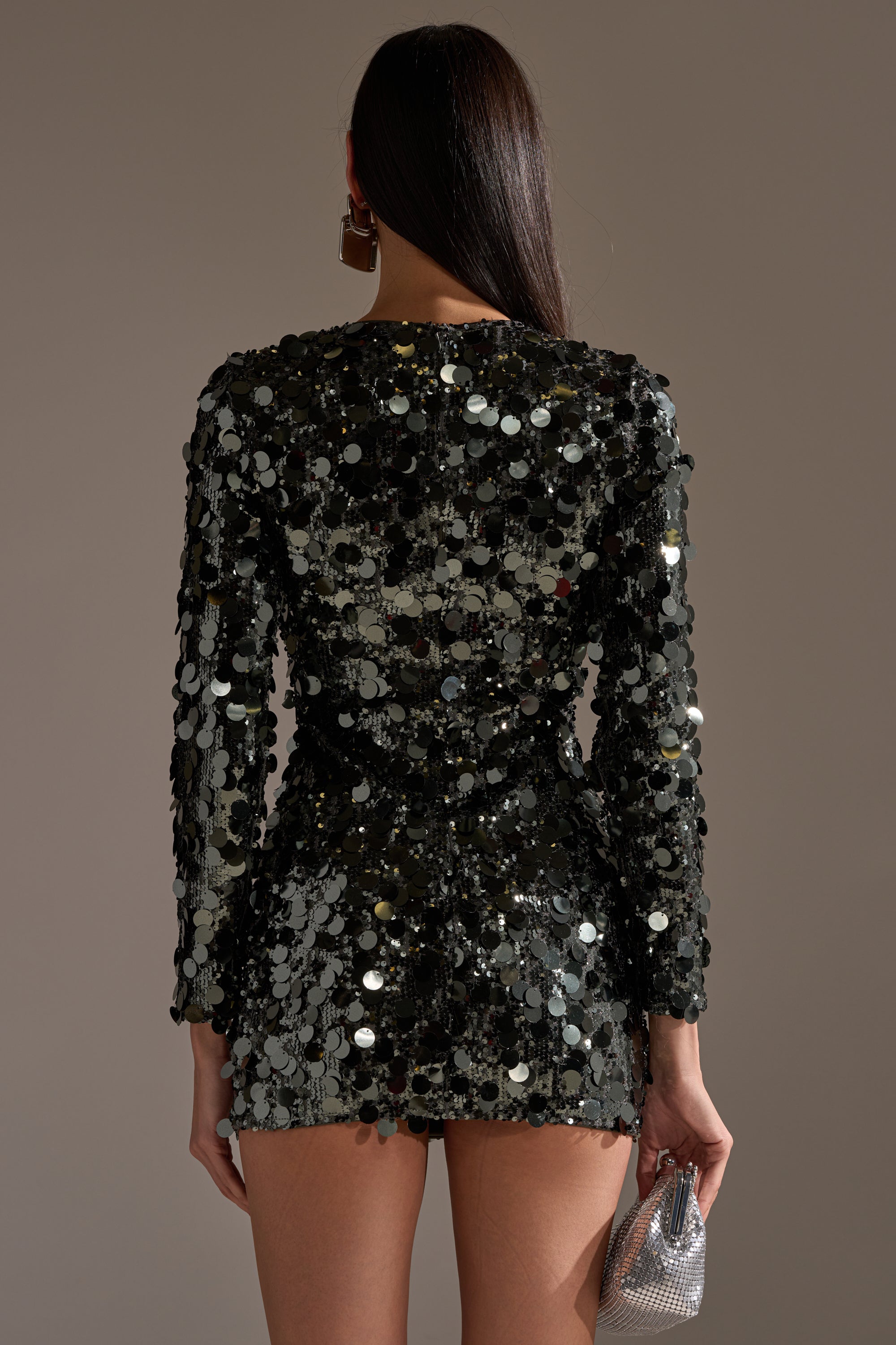 A person with long dark hair is seen from behind, wearing the SHE'S ALL THAT SEQUIN MINI DRESS IN OLIVE adorned with large reflective discs and holding a small, sparkling silver clutch—perfect for a chic party dress look.