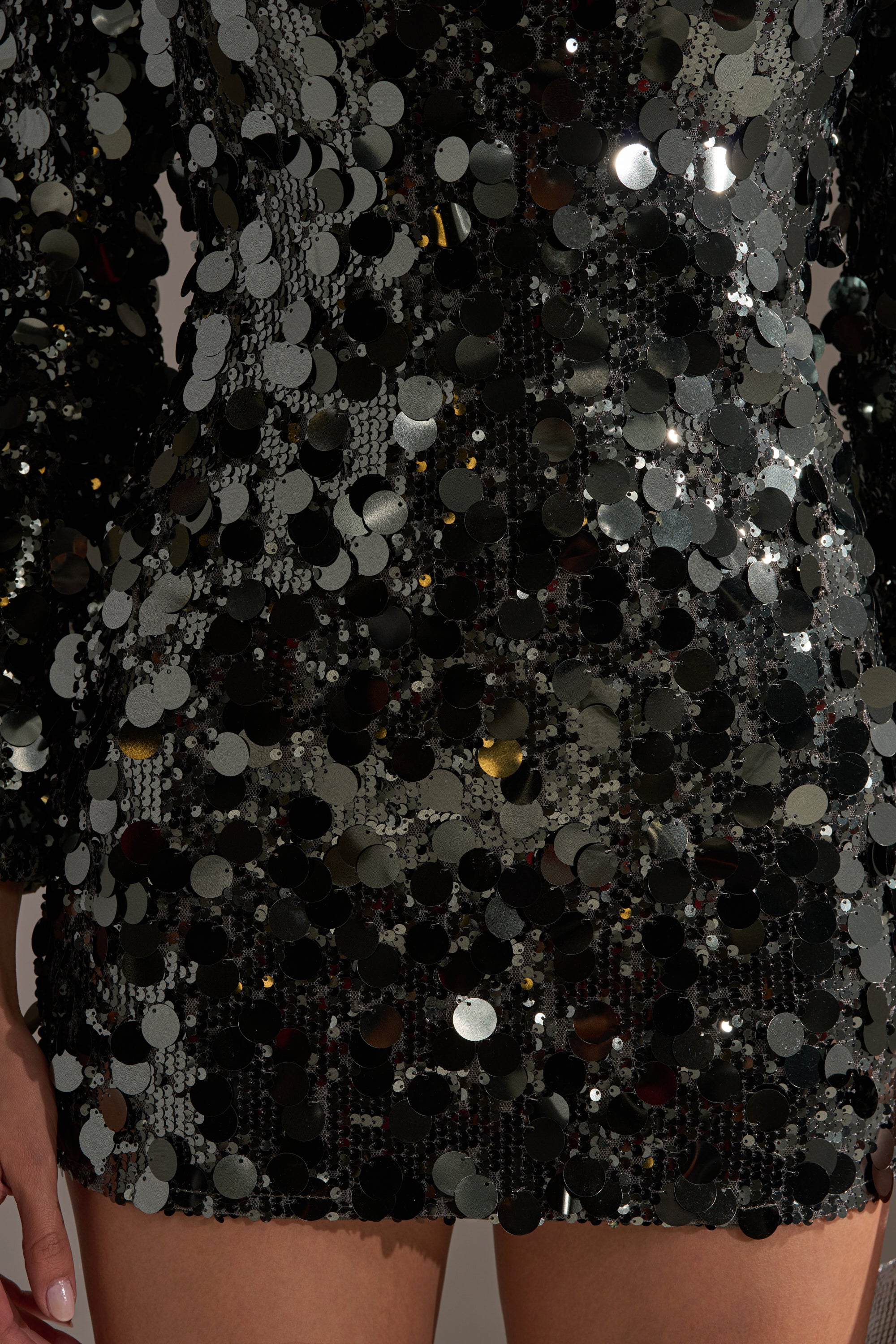 Close-up of a person wearing the SHE'S ALL THAT SEQUIN MINI DRESS IN OLIVE, featuring large, shiny circular sequins that reflect light. Hands rest at their sides and legs are visible from mid-thigh down—ideal for a standout party look.
