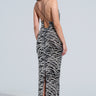 A woman stands facing away from the camera, adorned in the MEET ME AT MIDNIGHT SEQUIN AND MESH MAXI DRESS, featuring a zebra-inspired pattern with striking black and silver details. Her long dark hair flows down as she elegantly completes the ensemble with stylish black high heels against a simple, light-colored background.