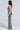 A woman stands in profile against a light background, wearing the MEET ME AT MIDNIGHT SEQUIN AND MESH MAXI DRESS. The dress features zebra-inspired black and silver sequin stripes with thin straps and an open back. She has long, wavy hair and is wearing high heels.
