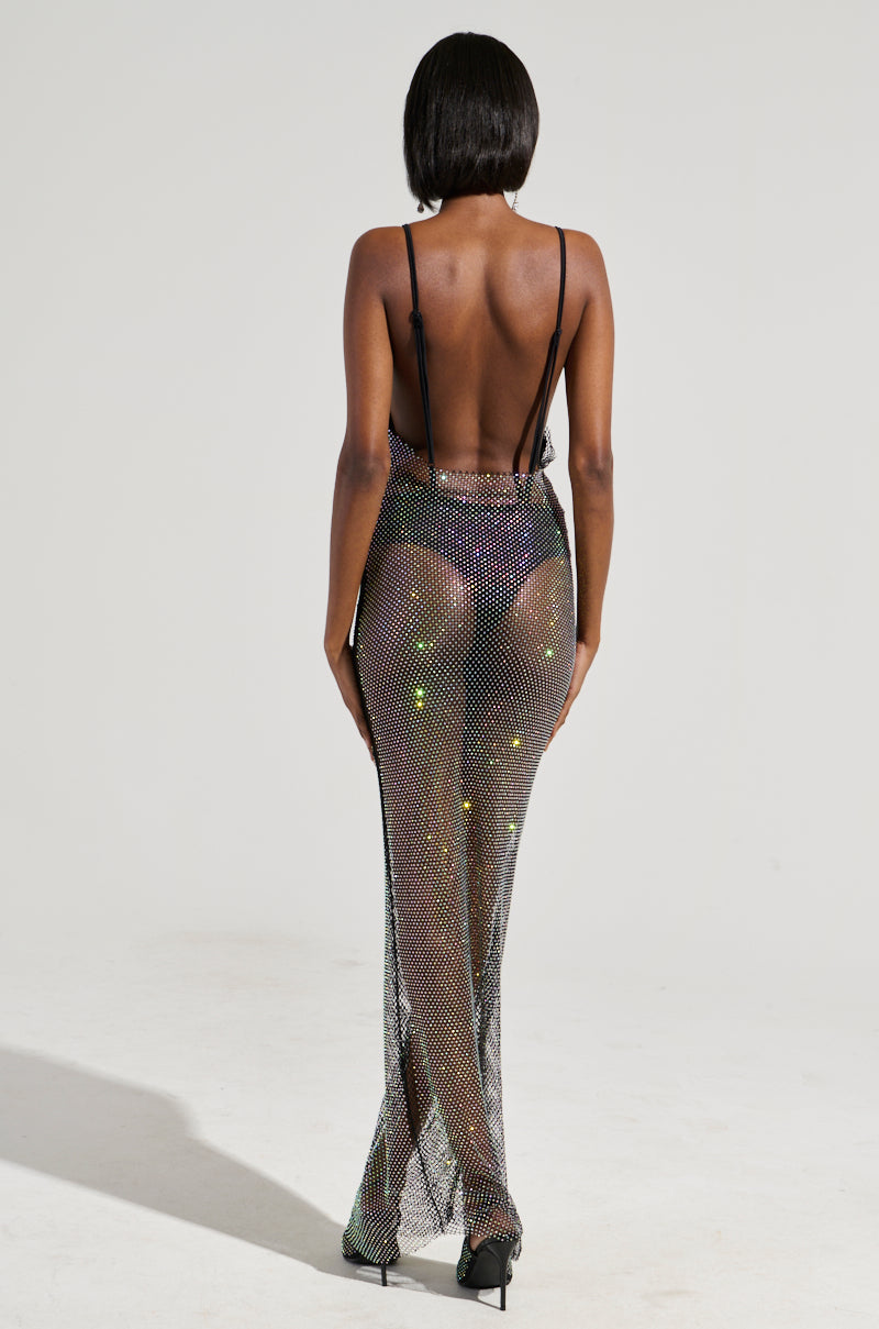 A person stands with their back to the camera, dressed in the Actin Brand New Rhinestone Mesh Maxi Dress paired with black high heels. The dress showcases a plunging back and delicate straps, elegantly revealing a black undergarment against a plain, light-colored background.