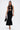 A woman wearing the NOT TOO CASUAL KNIT MAXI DRESS IN BLACK, featuring a sleeveless design, front cutout, and flared hem, stands against a white background with a black handbag and strappy heels.