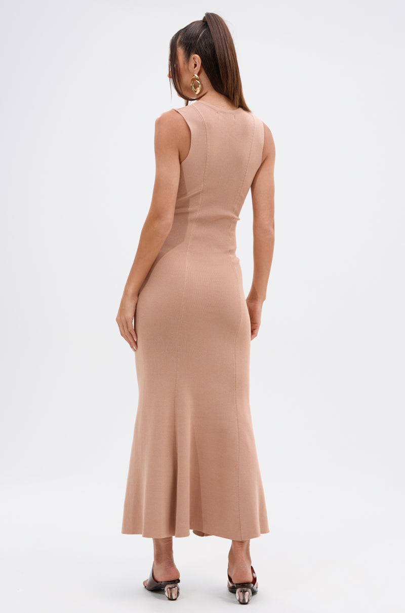 A woman with long brown hair in a ponytail wears the NOT TOO CASUAL KNIT MAXI DRESS IN BEIGE and heeled sandals, facing away from the camera against a plain white background.