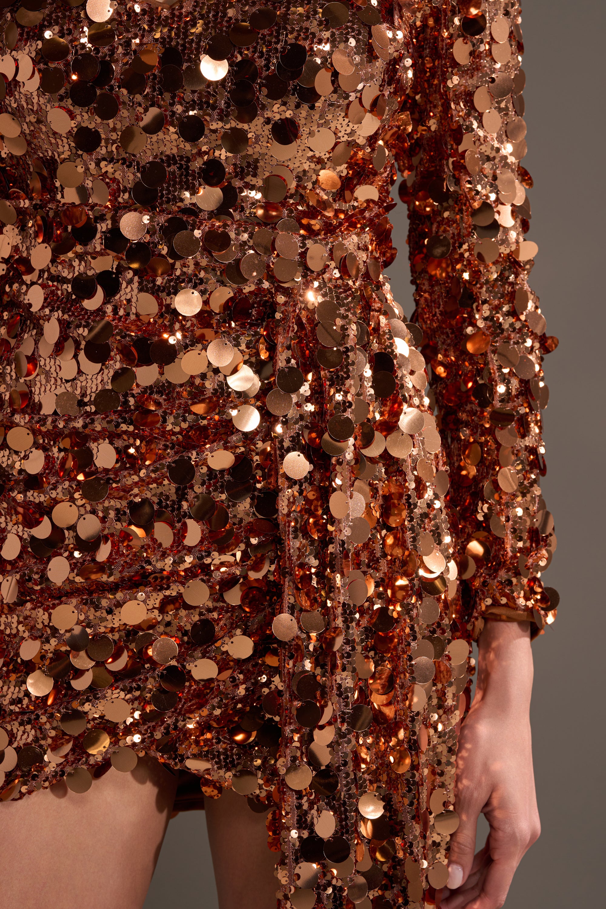 Close-up of a person wearing the DANCE THE NIGHT AWAY SEQUIN MINI DRESS, featuring bronze sequins, long sleeves, and a ruched design—perfect for evening events—set against a plain gray background.