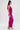 A woman stands in profile wearing the ALWAYS ON THE MOVE RUCHED MIDI DRESS in bright pink mesh, paired with silver high-heeled sandals. Her long brown hair is styled half-up, half-down as she poses against a plain white background.