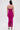 A woman with long brown hair faces away, wearing the ALWAYS ON THE MOVE RUCHED MIDI DRESS in bright pink stretchy mesh with back zipper and open hem, paired with white ankle-strap high-heeled sandals.