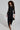 A person with curly hair wears the BUTTON UP BUTTER CUP COLLARED MAXI DRESS in black, featuring gold buttons and sheer mesh sleeves. They carry a handbag with ornate detailing and wear leopard-print shoes, standing against a plain background.