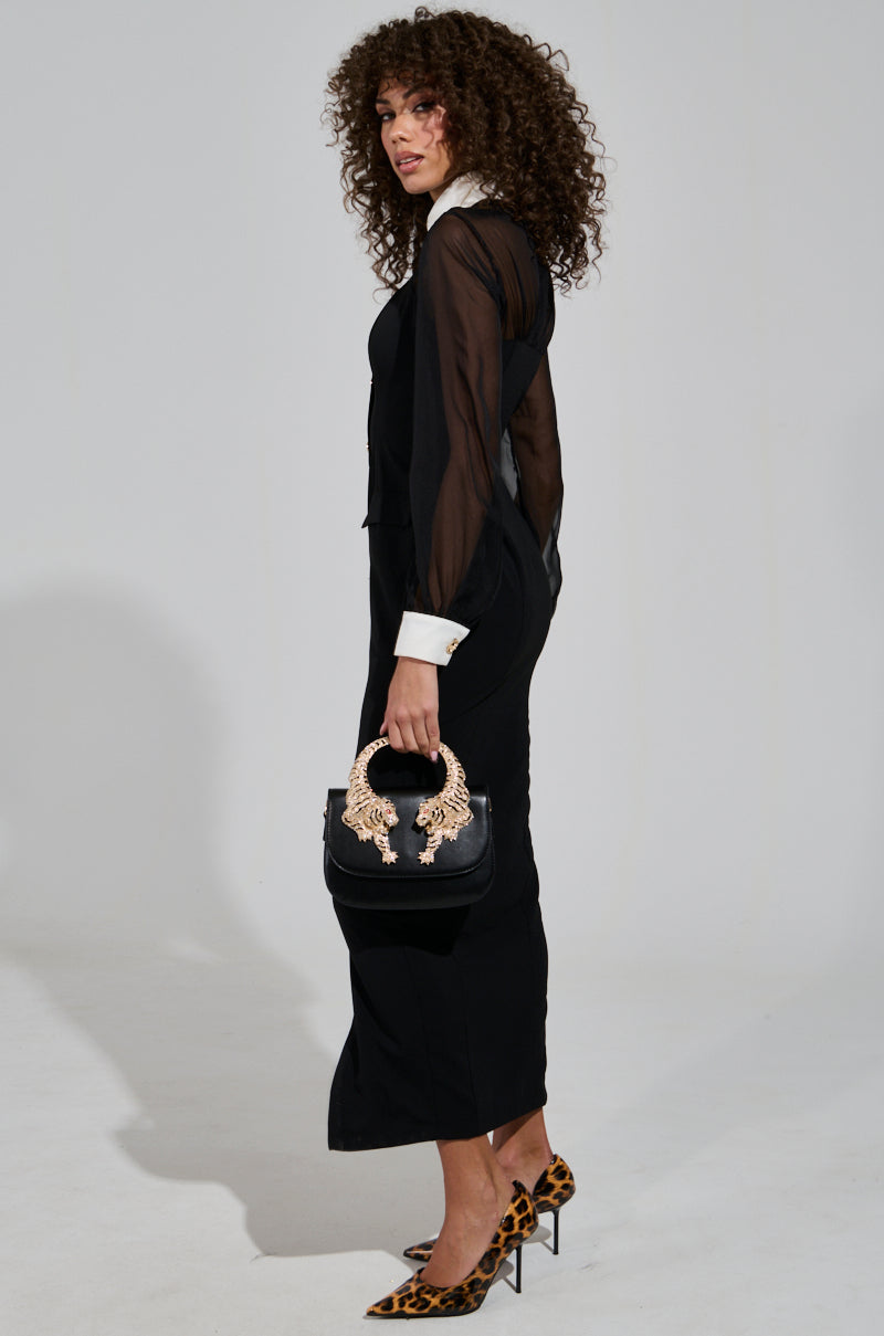 A woman with curly hair stands in profile against a plain background, wearing the Button Up Butter Cup Collared Maxi Dress, which features a sheer mesh bodice. She completes her look with leopard-print heels and holds a black handbag adorned with decorative handles.