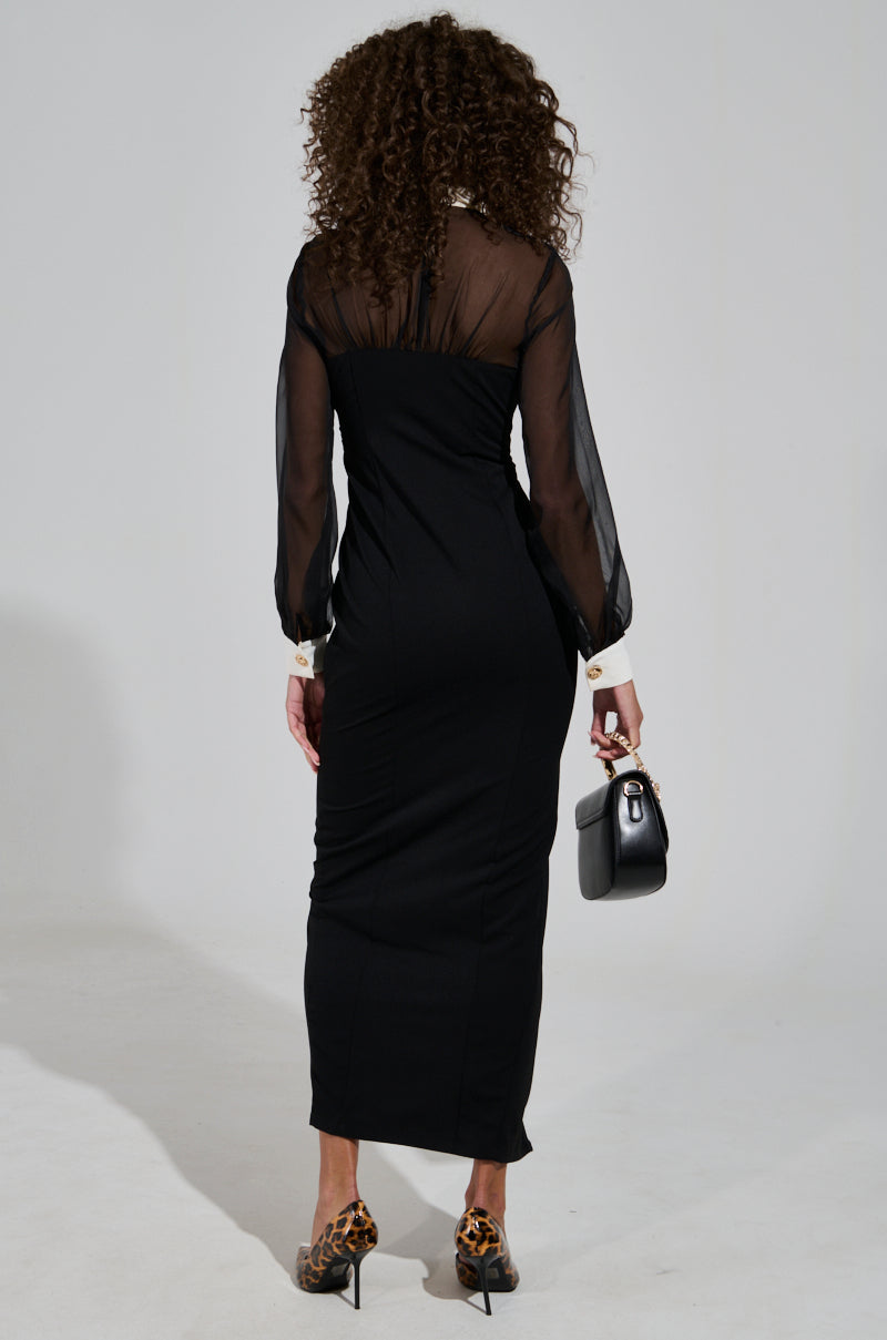 A person with curly hair stands with their back to the camera, wearing the Button Up Butter Cup Collared Maxi Dress in black and animal print high heels. The dress features sheer mesh sleeves, perfectly complementing the small black handbag they hold.