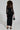 A person with curly hair stands with their back to the camera, wearing the Button Up Butter Cup Collared Maxi Dress in black and animal print high heels. The dress features sheer mesh sleeves, perfectly complementing the small black handbag they hold.
