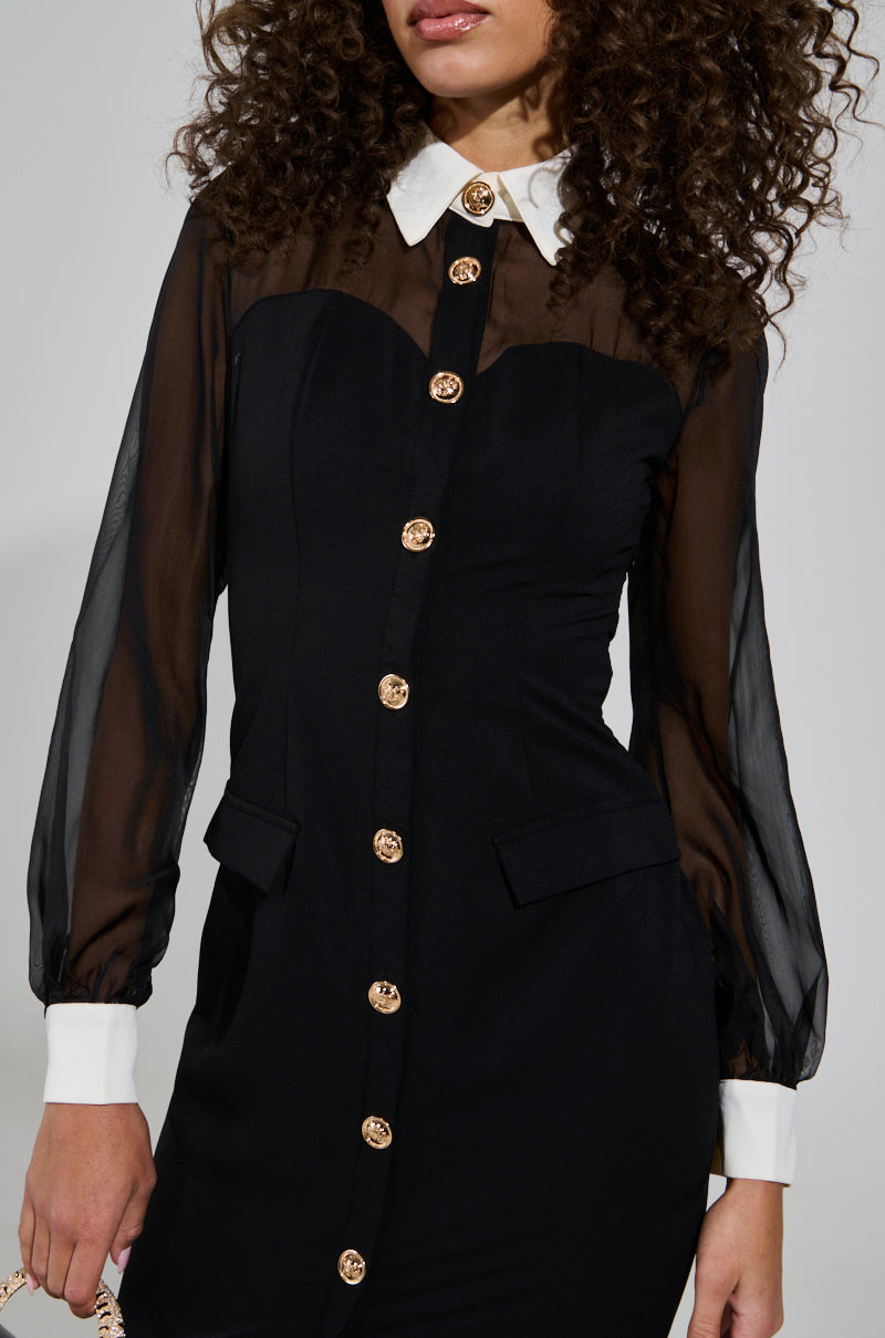 A woman wearing the BUTTON UP BUTTER CUP COLLARED MAXI DRESS, featuring sheer sleeves, a white collar, and gold buttons. The dress is designed with pocket flaps and a fitted silhouette. Her curly hair complements the look as she holds a bag against the plain backdrop.