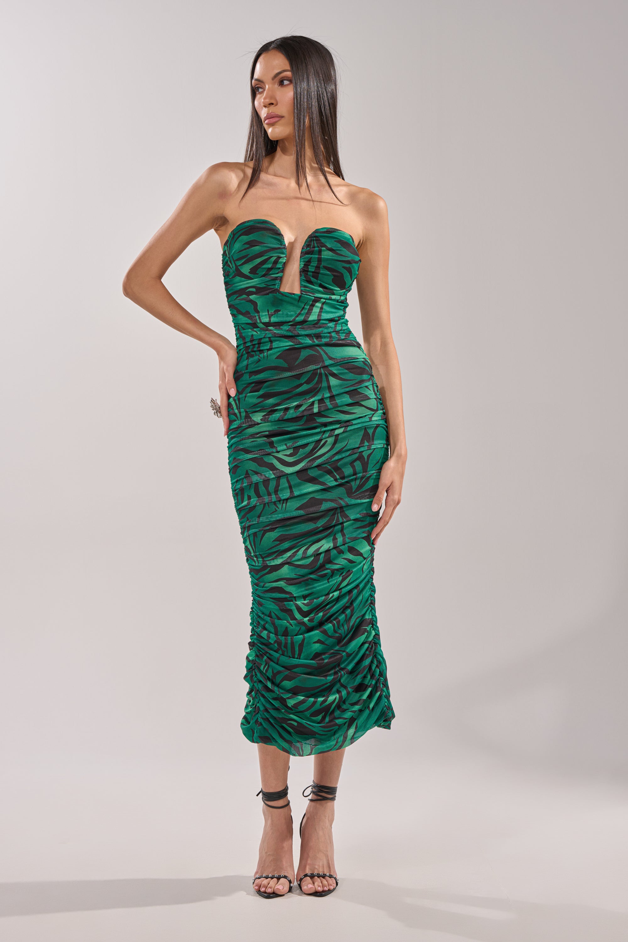 A woman wears the DAY OUT STRAPLESS PRINT MIDI DRESS, featuring a strapless sweetheart neckline and green zebra print with ruched details, paired with black high-heeled sandals. She stands in a neutral studio setting. 0