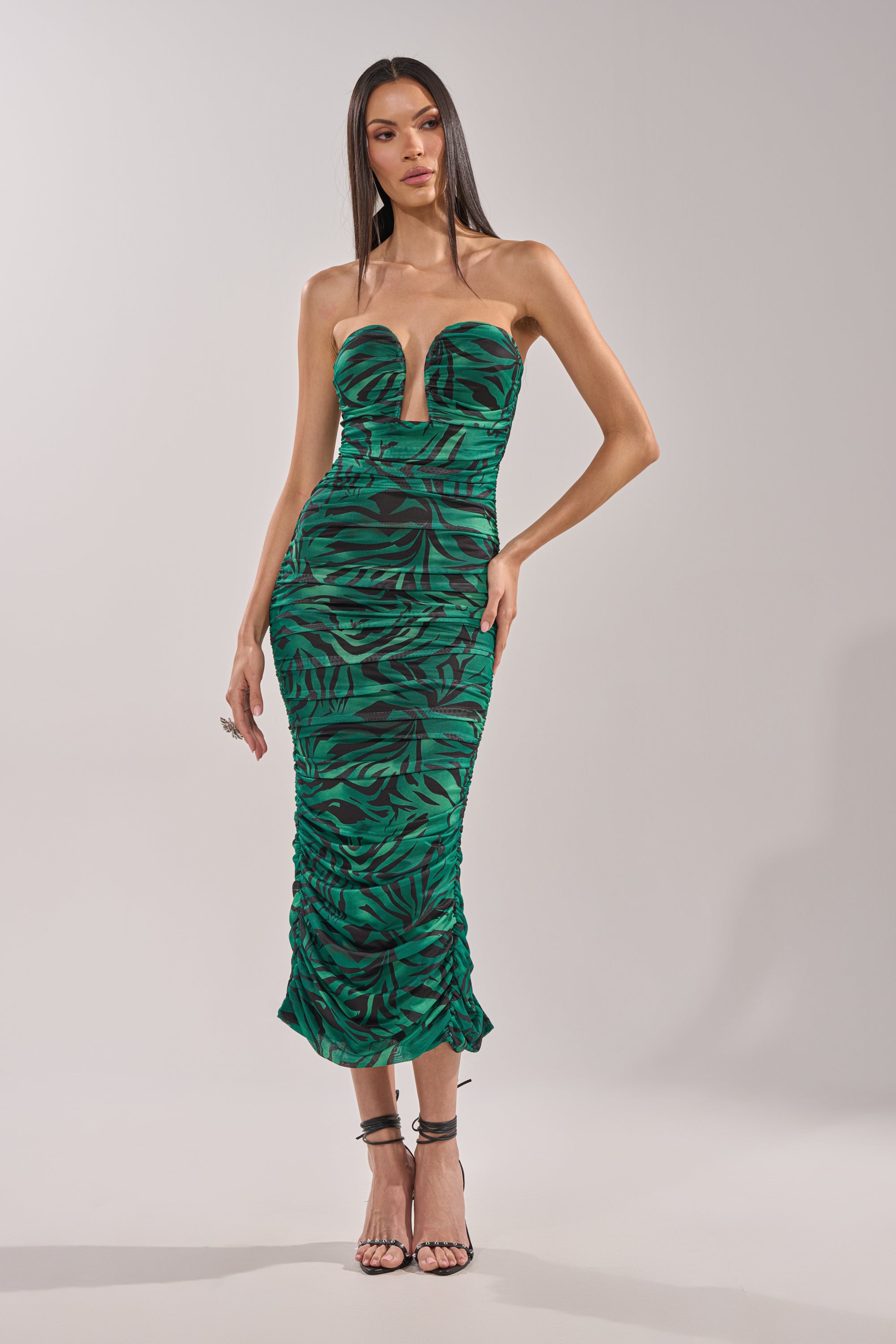 A woman models the DAY OUT STRAPLESS PRINT MIDI DRESS—a strapless, form-fitting green zebra print midi dress with a bold black leaf pattern and deep V neckline—paired with high-heeled sandals against a plain light background. 1