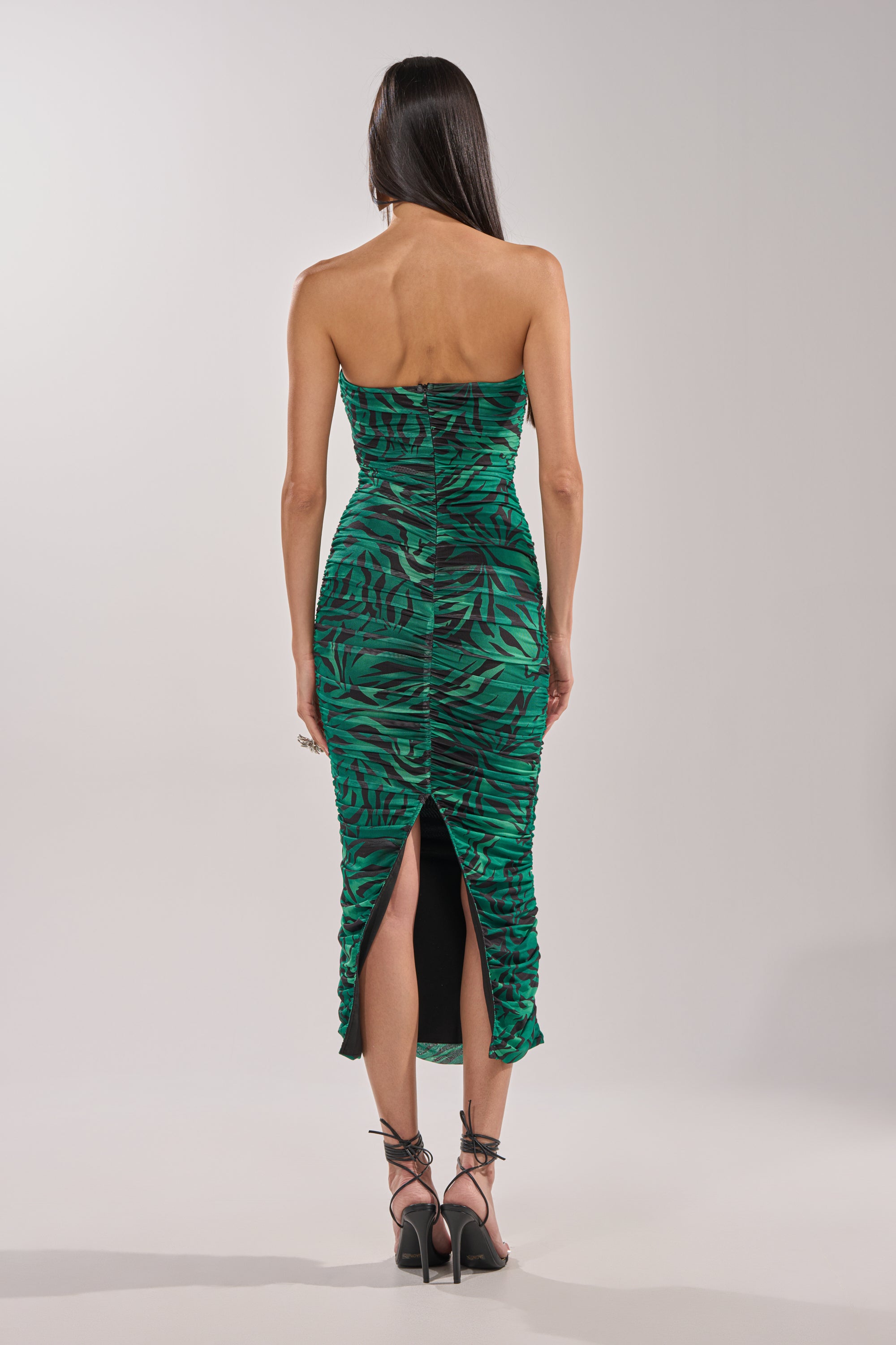 A woman with long dark hair models the DAY OUT STRAPLESS PRINT MIDI DRESS, featuring a green zebra print, strapless sweetheart neckline, ruched back, and back slit. She pairs it with strappy heels and stands facing away from the camera. 4