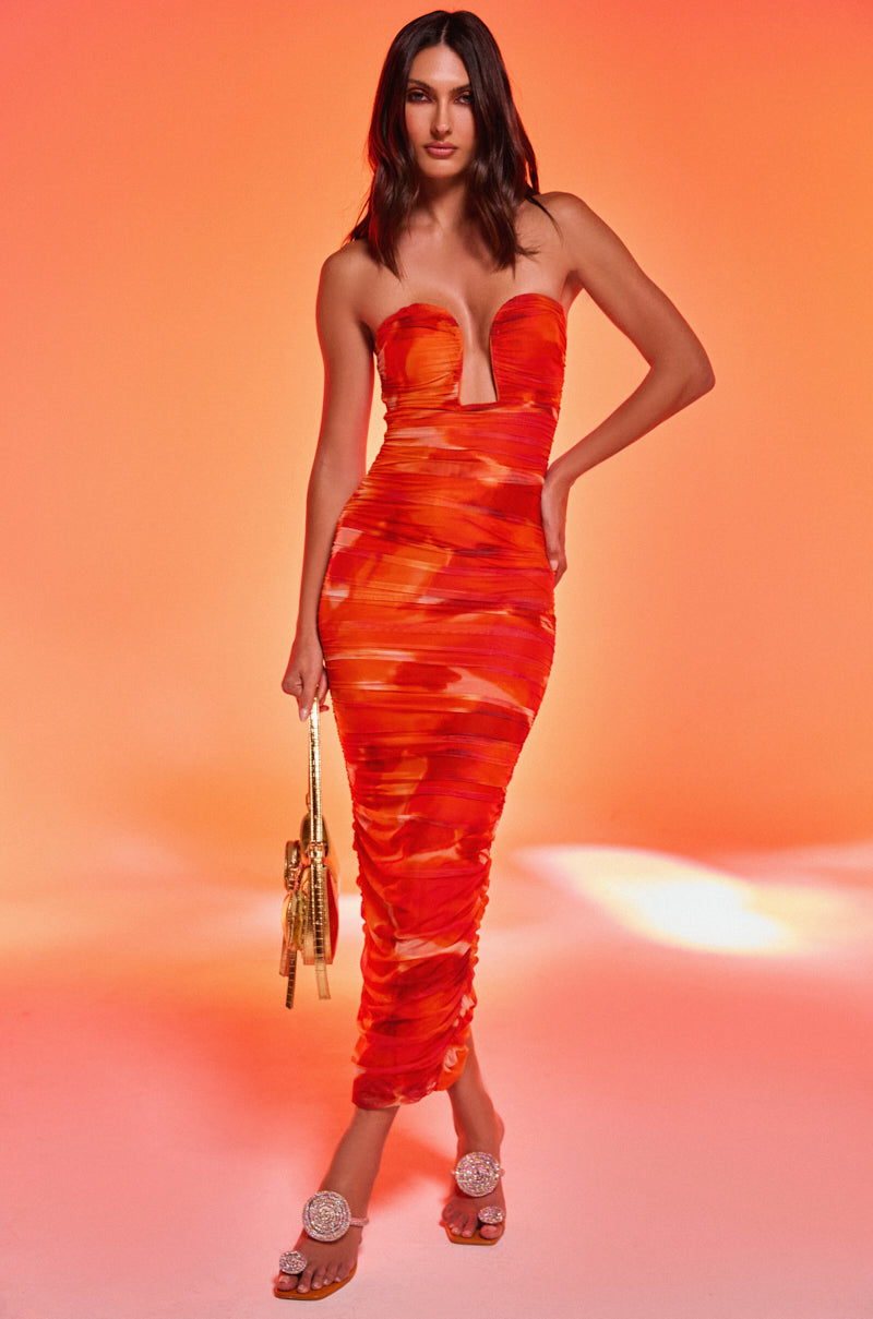A woman poses against an orange gradient background, wearing the LOTS OF LOVE OMBRE MIDI DRESS with a strapless neckline in vibrant orange and red. She accessorizes with a gold handbag and sparkling embellished heeled sandals.