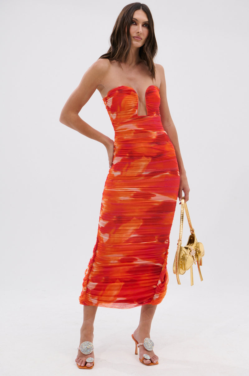 A woman poses in the LOTS OF LOVE OMBRE MIDI DRESS, showcasing its strapless neckline and deep V cut. She completes her look with a small gold handbag and orange heeled sandals adorned with bold round silver accents.