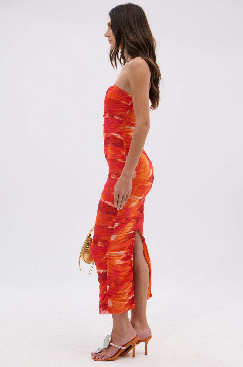 Woman standing sideways in the LOTS OF LOVE OMBRE MIDI DRESS—a fitted, strapless red and orange ombre midi dress with a side slit—holding a small handbag and wearing heeled sandals against a plain white background.