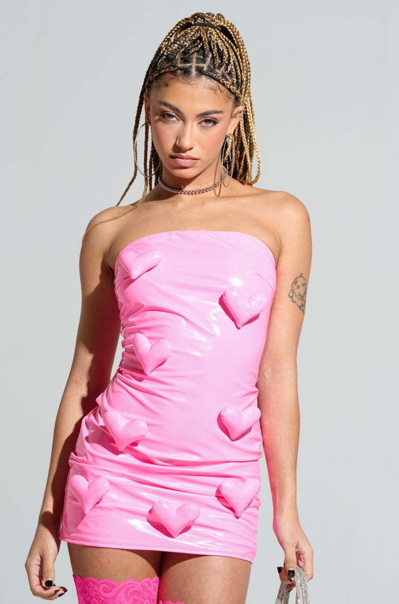 A person with braided hair wears the Barbie Dreams Faux Leather Mini Dress in pink, a strapless bodycon fit adorned with 3D heart shapes, paired with matching pink thigh-high stockings against a plain gray background.